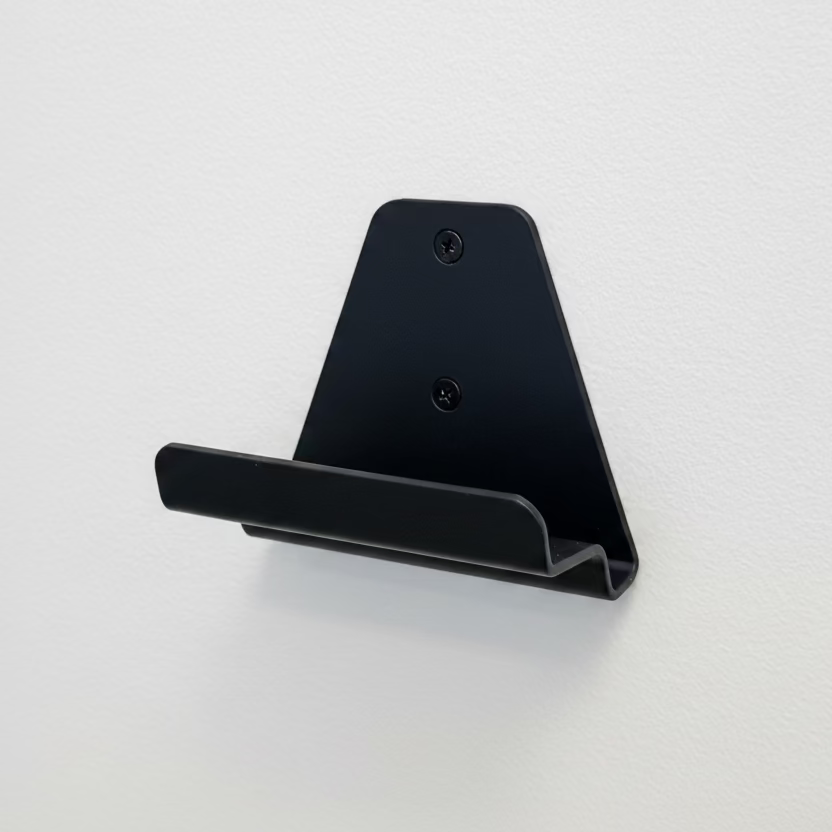 A matte black metal wall-mounted shelf bracket with two screw holes, shown mounted on a textured white wall.